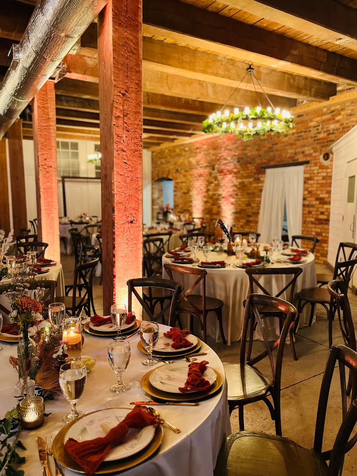 Indoor reception with round tables, rust napkins, gold chargers, and a greenery chandelier against brick walls